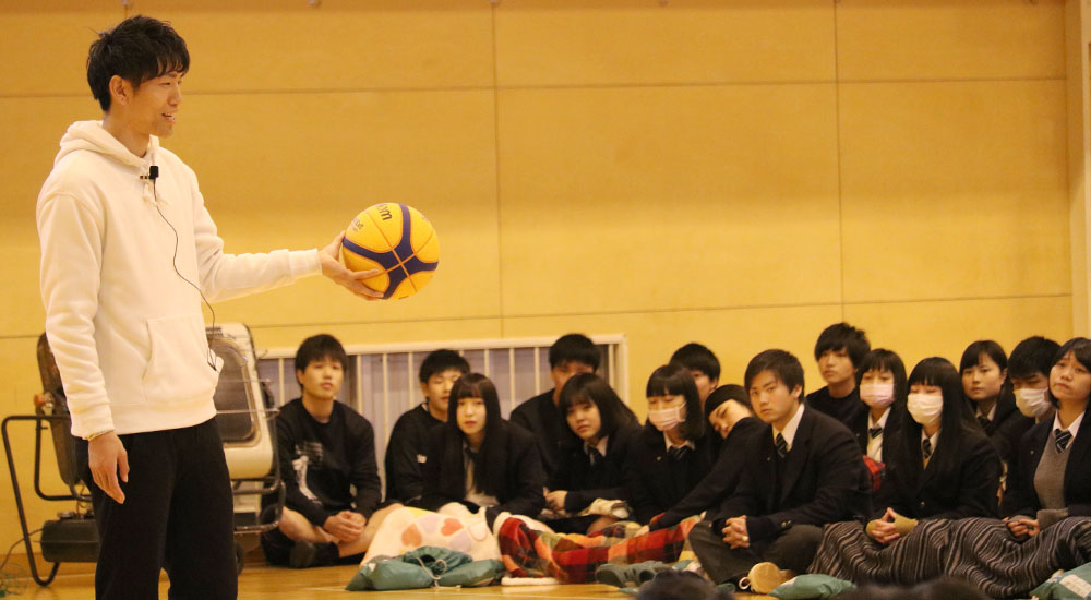 手にバスケットボールを持って高校生相手に講演を行う飯島康夫